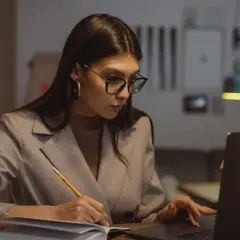 Kobieta w okularach pracuje przy laptopie, notując coś w zeszycie. Zastanawia się, ile maksymalnie nadgodzin w miesiącu może przepracować.