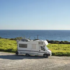 Camping-car blanc garé sur une aire gratuite en bord de mer en Bretagne. L'océan bleu s'étend à l'horizon, avec des champs verdoyants.