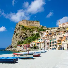 Vue d'une ville côtière italienne avec un château sur une falaise, des maisons colorées et des bateaux sur une plage.