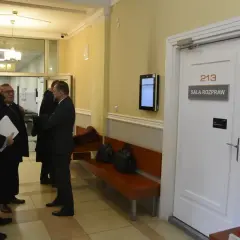 Trzech mężczyzn w togach prawniczych rozmawia przed drzwiami sali rozpraw nr 213.