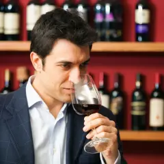 Mężczyzna w garniturze wącha czerwone wino w kieliszku. To degustator wina, który ocenia jego aromat.