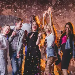 Grupa młodych ludzi świętuje, sypiąc konfetti. Ich stroje to świetna inspiracja, jak się ubrać do klubu: sukienki, jeansy, marynarki.