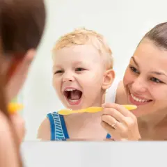 Mama uczy malucha, jak nauczyć dziecko myć zęby. Uśmiechnięty chłopiec z otwartymi ustami i żółtą szczoteczką.