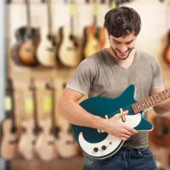 Uśmiechnięty młody mężczyzna trzyma turkusową gitarę elektryczną, idealną jako gitara dla początkujących. W tle widać rząd gitar.