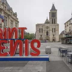Duże czerwone litery "SAINT DENIS" na tle bazyliki. Choć nazwa sugeruje niebezpieczne miejsca, plac wydaje się spokojny.