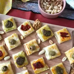 Rustici con pasta sfoglia, quadrati dorati con olive, pesto e pomodori secchi, sparsi su un tagliere con frutta secca.