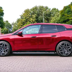 Błyszczący, czerwony BMW iX stoi na brukowanym placu, otoczony zielenią.