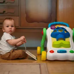 Małe dziecko siedzi obok kolorowego chodzika. Czy chodzik jest dobry dla dziecka? To pytanie nurtuje wielu rodziców.