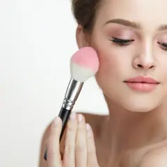 Mujer aplicando rubor en sus mejillas con una brocha. El tono rosado del rubor realza su piel, creando un look natural y radiante.