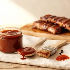 Sauce barbecue maison, prête à être appliquée sur des côtes grillées. Une cuillère déborde de sauce, un pinceau attend.