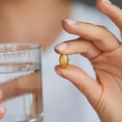 Dłoń trzyma kapsułkę omega 3 i szklankę wody. Kiedy brać omega 3? Najlepiej z posiłkiem.