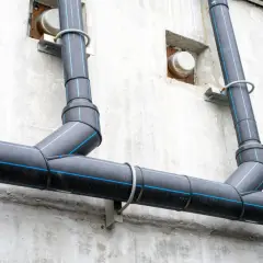Czarny pion kanalizacyjny z niebieskimi paskami biegnie wzdłuż szarej, betonowej ściany, przymocowany do niej metalowymi uchwytami.