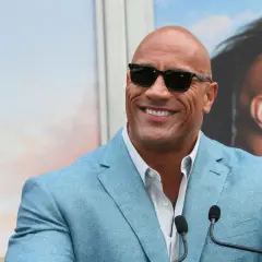 Dwayne "The Rock" Johnson, być może najbogatszy aktor na świecie, w błękitnej marynarce i okularach przeciwsłonecznych, uśmiecha się do kamery.