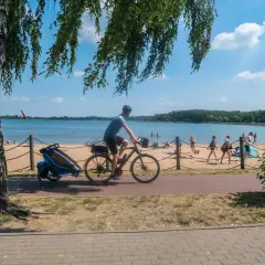 Rowerzysta z przyczepką na plaży nad jeziorem, w tle ludzie odpoczywający na piasku. Trasa Pogoria 3.