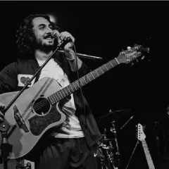 Un postino cantante con chitarra e microfono sul palco, circondato da strumenti musicali.