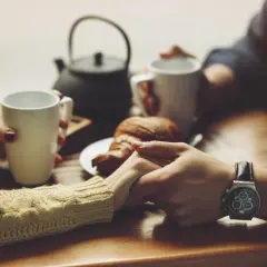 Dłonie trzymające kubki z kawą, croissant na talerzyku. W tle czajniczek. To idealny moment na szczere komplementy dla kobiety.