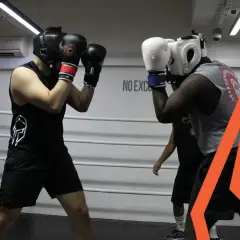 Two boxers spar in a ring, practicing their boxing defense. One boxer wears a black tank top and headgear, the other a grey tank top and white headgear.