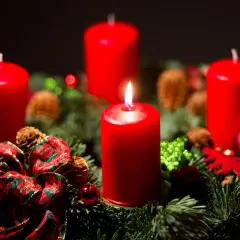 Ein Adventskranz mit vier roten Kerzen, Tannenzweigen, Zapfen und Schleifen. Die erste Kerze brennt und symbolisiert, warum man Advent feiert: die Vorfreude auf Weihnachten.