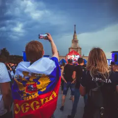 Młody kibic z flagą Rosji, wspierający klub piłkarski z Moskwy, filmuje telefonem wydarzenie przed monumentalnym budynkiem.