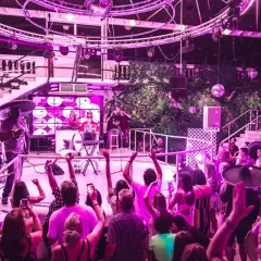 Festa a tema anni '90 a Ibiza, con DJ, performer con sombrero e folla in delirio sotto le luci stroboscopiche.
