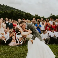 Para młoda całuje się na tle rodziny i przyjaciół. To piękny moment z planu dnia ślubu, pełen radości i miłości.