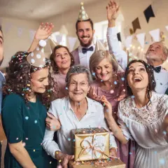 Radosna rodzina świętuje 100 rocznicę ślubu. Wszyscy w czapkach, sypią się konfetti. Babcia trzyma prezent.