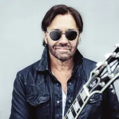 Al Di Meola w czarnej skórzanej kurtce i okularach przeciwsłonecznych, trzymający gitarę.