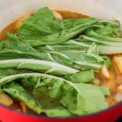 Verduras frescas, como acelgas, se cocinan en una olla roja con trozos de patata en un caldo sabroso.