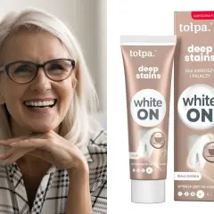 Uśmiechnięta kobieta z jasnymi włosami i okularami. Obok tubka i pudełko pasty do zębów Tołpa Deep Stains White ON – jaka pasta wybielająca dla kawoszy i palaczy.