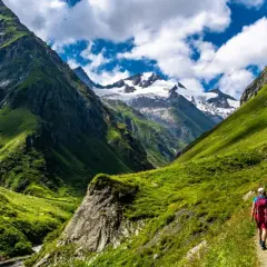 Hikers on a trail enjoying alpine hiking amidst lush green mountains and snow-capped peaks under a cloudy blue sky.