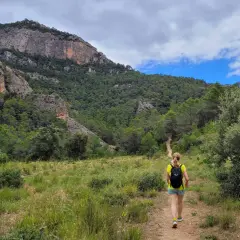 Senderista camina por un sendero en los **estrets d'Arnes**, rodeada de pinos y montañas rocosas bajo un cielo nublado.