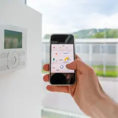Control de la automatización de casas desde un smartphone. Se ve un termostato en la pared y una mano sujetando un teléfono con una app de control.