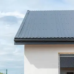 Szary dach z blachy falistej, z rynną, o kącie nachylenia dachu 15 stopni, nad białym murem domu.