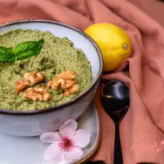 Un delicioso pesto vegano en un cuenco, adornado con hojas de albahaca y nueces. Junto a un limón y una flor rosa.