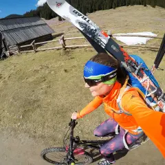 Kobieta na rowerze z nartami na plecach w górach. To może być jej sposób na roztrenowanie po sezonie.