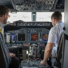 Dwóch pilotów samolotu w kokpicie. Jeden z nich, pilot, patrzy na instrumenty, drugi obsługuje sterowanie.