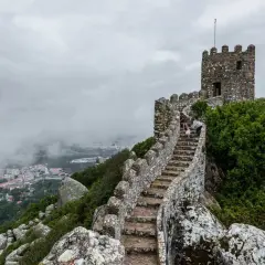 Escaleras de piedra ascienden hacia una torre en el **castillo de Sintra**, con vistas a la ciudad envuelta en niebla.