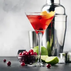 Elegancki Cosmopolitan przepis: czerwony drink w kieliszku, obok żurawina, limonka i shaker. Idealny na wieczór.
