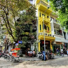 Ulica w Hanoi z żółtą kamienicą, rikszą i skuterami. W tle widać sklepy i restauracje, gdzie można wymienić walutę w Hanoi.