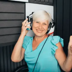 Uśmiechnięta kobieta w słuchawkach podczas badania audiometrii tonalnej. Pokazuje kciuk w górę, co oznacza pozytywny wynik.