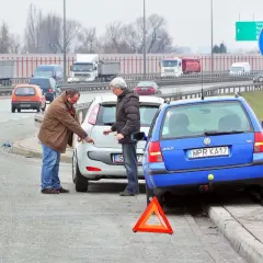 Dwóch mężczyzn dyskutuje przy uszkodzonych samochodach po kolizji. Trójkąt ostrzegawczy sugeruje, że może być spowodowanie kolizji mandat.