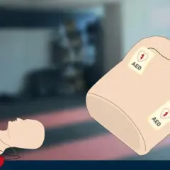 Ilustracja przedstawia manekina do resuscytacji z przyklejonymi elektrodami AED oraz czerwony defibrylator AED.