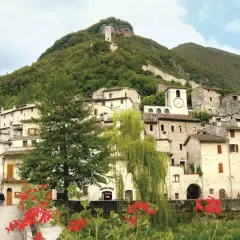 Antico borgo arroccato su un colle verdeggiante, uno dei paesi della Valnerina, con case in pietra, un campanile con orologio e una torre medievale in cima.