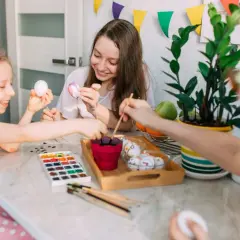 Dzieci malują pisanki przy stole, w tle kolorowe chorągiewki.
