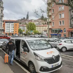 Un homme charge une valise orange dans un bus compostelle blanc. La ville s'étend en arrière-plan.