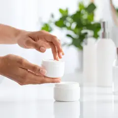 Dłonie nakładają krem z PEG-60 hydrogenated castor oil na twarz. W tle kosmetyki i rośliny.