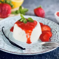 Cremige Panna Cotta mit Erdbeersauce, frischen Erdbeeren, Minze und einer Vanilleschote auf einem Teller.