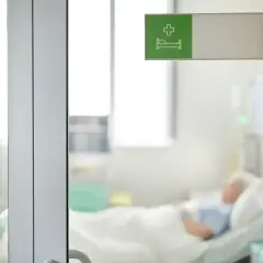 Szpitalne drzwi uchylone, w tle pacjent na łóżku. Czasem wybudzanie ze śpiączki farmakologicznej trwa długo.