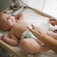 Radosna pielęgnacja niemowlaka chłopca. Mama zmienia pieluszkę, maluch uśmiecha się, leżąc na przewijaku wśród zabawek.
