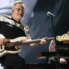 Muzyk gra na białej gitarze basowej, tworząc swoje opus magnum. W tle klawiszowiec pochyla się nad instrumentem.